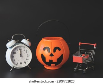 Front view of  Halloween pumpkin bucket  with shopping cart and white vintage alarm clock 12 pm. isolated  on black  background with copy space. Halloween holiday midnight shopping  concept.