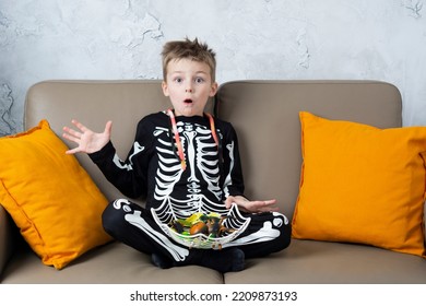 Happy shocked shouts halloween boy having fun at home eat scary candy and looking at camera wearing Halloween costume skeleton on home background.