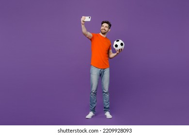 Full body jonge fan man hij draagt ​​oranje t-shirt opvrolijken ondersteuning voetbal sport team houden voetbal kijken tv live stream doen selfie geschoten op mobiele telefoon geïsoleerd op effen donkere paarse achtergrond