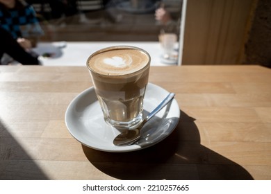 Flat white coffee in clear transparent glass and white plate lay on rough wooden table counter beside windows of coffee shop and blur background of outdoor seats and people outside coffee shop.