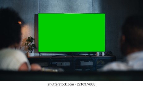 Achteraanzicht van het gelukkig getrouwde zwarte stel dat popcorn eet op de bank en tv kijkt met een groen scherm Mock Up Display in de woonkamer van de loft. Onscherp paar close-up shot 's nachts.