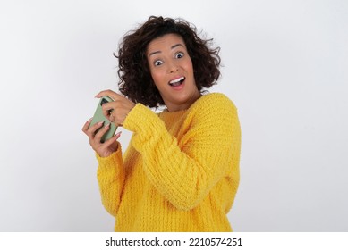 Wanita cantik berambut cokelat muda yang cantik dan ceria dengan rambut pendek keriting mengenakan sweter kuning di atas dinding putih menggunakan gadget bermain game jaringan