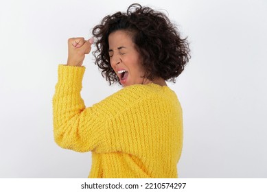 Potret wanita muda berambut cokelat cantik yang lucu dengan rambut pendek keriting mengenakan sweter kuning di atas dinding putih berteriak ya angkat tangan merayakan kompetisi permainan kemenangan