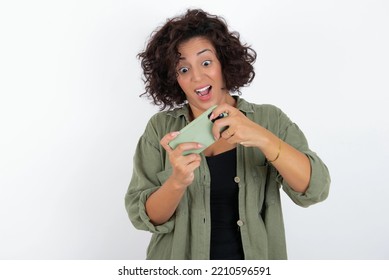 Potret seorang wanita muda cantik yang bersemangat dengan rambut pendek keriting mengenakan kaus hijau di atas dinding putih bermain game di ponsel.