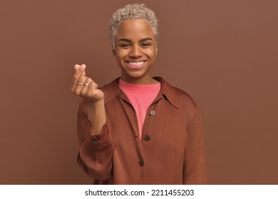 Young beautiful blond African American woman smile and makes Korean love sign, shapes heart with her fingers expresses her love and sympathy, stand over brown background. Hand gesture concept.