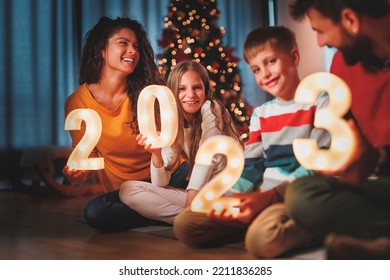 Orang tua merayakan Malam Tahun Baru di rumah bersama anak-anak, duduk di dekat pohon Natal, memegang angka iluminatif 2023 yang mewakili Tahun Baru yang akan datang