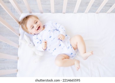 bambino biondo che ride sdraiato sulla schiena in una culla in una stanza di casa con biancheria da letto di cotone blu e bianco, sorridente al mattino, il concetto di beni e accessori per bambini