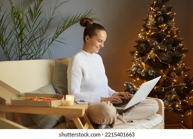 Portrait de côté d'une jolie jeune femme adulte souriante portant un pull blanc assise sur un canapé et travaillant sur un ordinateur portable, regardant l'écran de l'ordinateur portable, réalisant un projet en ligne pendant le réveillon du Nouvel An.