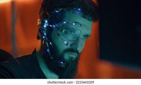 Un ragazzo dai capelli castani lavora al computer con luci al neon sullo sfondo. Stile cyberpunk. Indossa occhiali futuristici monoculari e un microfono. Sfondi cyberpunk e fantascientifici.