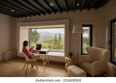 Femeia lucrează la laptop în timp ce stă lângă masă în fața ferestrei panoramice cu vedere minunată asupra munților. Vedere interioară largă. Conceptul de lucru la distanță și evadare în natură