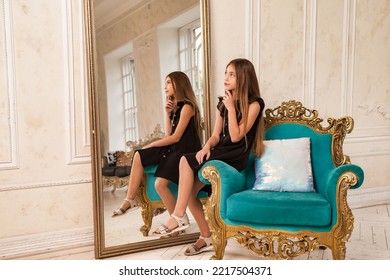 Teenager girl princess 10 year old with curly hair sits on armchair, fashion model in stylish black elegant dress in living room with mirror, looking away. Fashionable young lady actress. Copy space