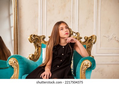 Teenager girl princess 10 year old with curly hair sitting on armchair, fashion model in stylish black elegant dress in living room, looking away. Fashionable young lady actress. Copy text space