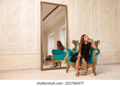 Teen girl princess 10 years old with curly hair sitting on armchair, fashion model in stylish black elegant dress in empty room with mirror, looking away. Fashionable young lady actress. Copy space