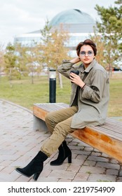 Elegante modelo asiático tocando el pelo corto mientras se sienta con café en un banco al aire libre. Hermosa joven con gafas y ropa verde posando y mirando hacia otro lado.