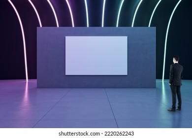 Vista trasera de un hombre de negocios mirando un afiche blanco en blanco con espacio para su logotipo o texto en una partición oscura en una sala abstracta con suelo de hormigón y lámparas iluminadas con neón en un fondo oscuro, maqueta