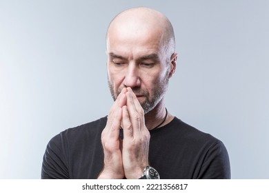 A serious man folded his hands in supplication. Bald brutal guy in a black shirt. Sadness, longing and request for support. Grey background. Close-up.