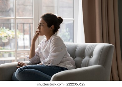 Sad upset young Indian woman thinking while sit on armchair and staring out window. Personal troubles and break up, goes through difficult life situation, search solution, ponders seated alone at home