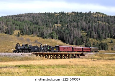 Twee historische stoomlocomotieven trekken een antieke houten passagierstrein over een spoorbrug in een schilderachtige weide met een bergbos op de achtergrond