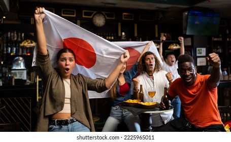 Excited young woman, Japan football team fan, spending time in bar with friends while watching game. People with state flag in pub.