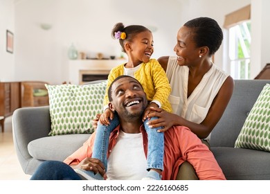 Niña alegre sentada en el hombro del padre mientras juega con la madre en casa. Familia negra feliz disfrutando el fin de semana en casa. Linda hijita sentada en el hombro de los padres y jugando con su madre.