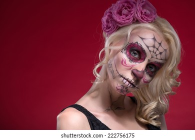 Chân dung cận cảnh của cô gái trẻ tóc vàng với khuôn mặt buồn với lớp trang điểm Calaveras và một bông hoa hồng trên tóc đang tò mò nhìn vào máy ảnh, tách biệt trên nền đỏ với vị trí sao chép