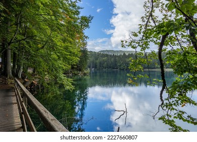 panoramic view from behind the trees on the secondary Fusine Lake with the green forest, the blue sky and the white clouds reflecting on the water surface in the afternoon in summer
