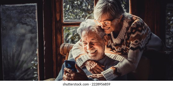 Happy senior old couple use mobile phone at home together to video call parents. Winter season indoor leisure activity for retired mature people. Woman hug mature man on the chair. Love and technology