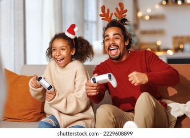 ครอบครัวแอฟริกันอเมริกันที่มีความสุข พ่อและลูกสาวหัวเราะและเล่นวิดีโอเกมด้วยกันโดยใช้คอนโซลวิดีโอเกมในช่วงวันหยุดคริสต์มาส