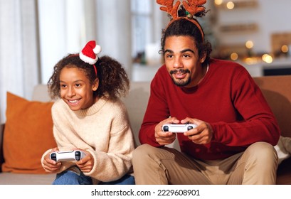 ครอบครัวแอฟริกันอเมริกันที่มีความสุข พ่อและลูกสาวหัวเราะและเล่นวิดีโอเกมด้วยกันโดยใช้คอนโซลวิดีโอเกมในช่วงวันหยุดคริสต์มาส