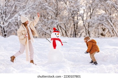 Gelukkige dolgelukkige familie moeder en zoontje spelen sneeuwballen buiten in de winter terwijl ze in de buurt van sneeuwpop staan, moeder en kind lachen, plezier hebben en spelen met sneeuw in het besneeuwde winterpark