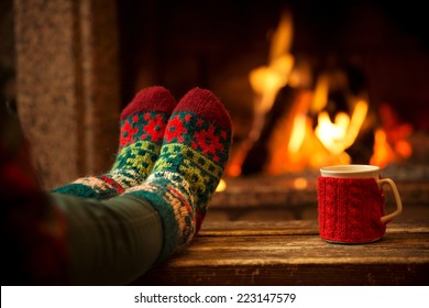 Voeten in wollen sokken bij de kersthaard. Vrouw ontspant bij warm vuur met een kop warme drank en warmt haar voeten op in wollen sokken. Close-up op voeten. Winter en kerstvakantie concept.