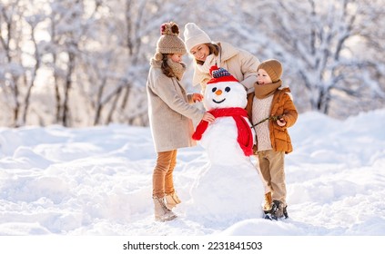 Young mom and two kids making snowman in winter park outdoors. Joyful little brother and sister in warm clothes happily helping mother decorate snow figure with carrot nose and hands with dry branches