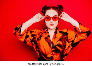 Cute girl on a red background. Red hair and tunnels in the ears, red dress and lips. Anime poses. A girl in orange sunglasses and two buns on her head looks at the camera.