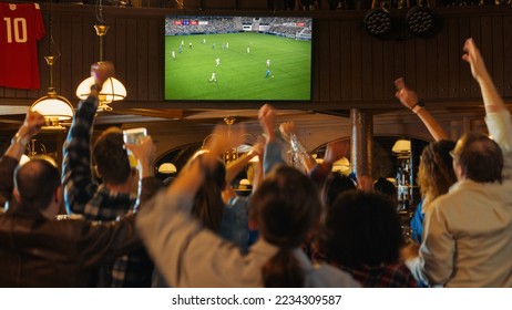 Grupo de aficionados al fútbol viendo un partido de fútbol en vivo en un bar deportivo. Personas de pie frente a un televisor, animando a su equipo. El jugador marca un gol y la multitud celebra ganar el campeonato.