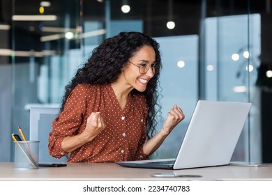 Hispanesch Geschäftsfra feiert den Erfolleg vum Victoire, Mataarbechterin mat gekrauselten Hoer am Büro liest gutt Neiegkeeten, benotzt e Laptop op der Aarbecht am Büro, hält d'Hand an d'Luucht an eng glécklech Triumphgest.