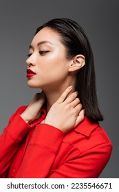 potret wanita asia berjaket merah berpose dengan tangan di leher terisolasi di abu-abu