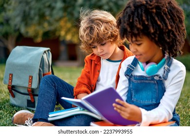 Adorable schoolchildren doing their homework together while sitting on the grass in the park. Back to school. African American girl with Caucasian cute boy communicate after school.
