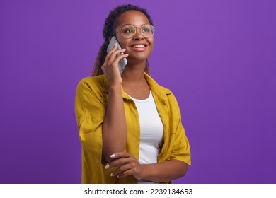 Fiatal pozitív etnikai afroamerikai nő szemüveges, mosolyogva felnéz, telefonál és flörtöl a barátjával vagy a hétköznapi stílusban öltözött munkatársával, pózol a lila stúdióban.