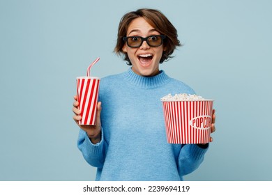 Mujer joven emocionada y sorprendida con gafas 3D mira una película mientras sostiene en la mano un vaso de refresco de cola, un cubo de agua con gas y palomitas de maíz en el cine aislado sobre un fondo azul cian claro pastel liso retrato de estudio