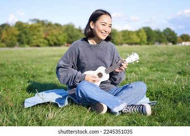 美しいアジアの女の子が公園に座って、ウクレレを弾きながら歌い、晴れた春の日に屋外でリラックスしています。