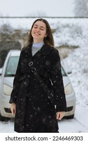 Ženska blizu avta. Sneženje in ženska. Lepa fotka blizu avta. Voznica. Dekle pozimi v plašču stoji blizu sivega avtomobila. Rjavolaska z avtomobilom. Dekle pozira blizu avtomobila. Avtomobil v naravi.