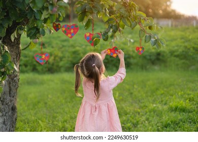 Baby Girl decorates apple tree branches with hearts postcards with multicolored puzzle pieces inside on day of autism awareness. Teach to be kind and tolerant from childhood