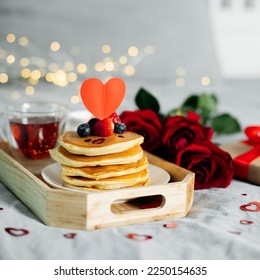 Valentine's day composition with breakfast, gift box and roses. Stack of delicious pancakes with berries and cup of hot moning tea on wooden tray.