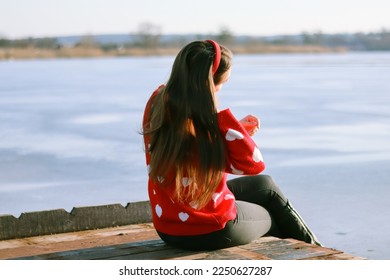Woman in motion.Woman in a sweater with hearts.Sits on the pier.Long legs.Loneliness.Resting near the lake.The wind blows her hair.Stylish photo.Retro and vintage.Bokeh and blur.Lonely.Valentine's Day