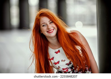 una chica pelirroja con una falda ligera; una chica muy alegre de pelo largo y rojo; labios pintados con lápiz labial rojo; se sienta en un banco y tiene buen humor; posa para un fotógrafo; sesion de fotos en la calle