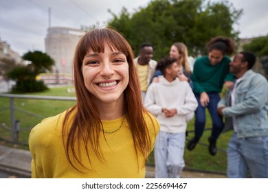 Portrait of a Young red-haired girl with wide smile and piercing look at the camera. Group of friends gathered in the background of the image. Colleagues hanging out in the city. Outdoors.