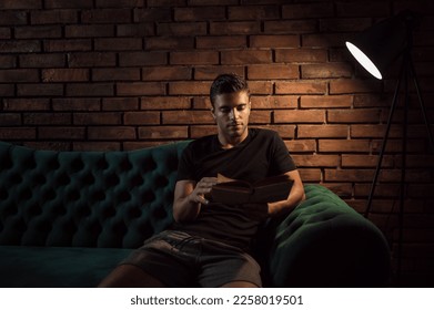 Portrait of young handsome man holding book, sitting on couch at home in living room, reading literature