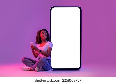 Mujer negra sonriente usando un teléfono móvil mientras está sentada cerca de un enorme teléfono inteligente en blanco con una pantalla blanca vacía, mujer afroamericana posando con luz de neón sobre un fondo de estudio morado, maqueta