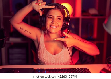 Wanita muda berambut pirang bermain video game sambil mengenakan headphone sambil tersenyum membuat bingkai dengan tangan dan jari dengan wajah bahagia. konsep kreativitas dan fotografi.