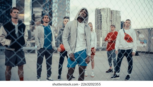 一位身穿连帽衫的英俊健壮黑人男子站在城市停车场屋顶的栅栏后面，摆出姿势，看着镜头。他身后站着一群不同的朋友